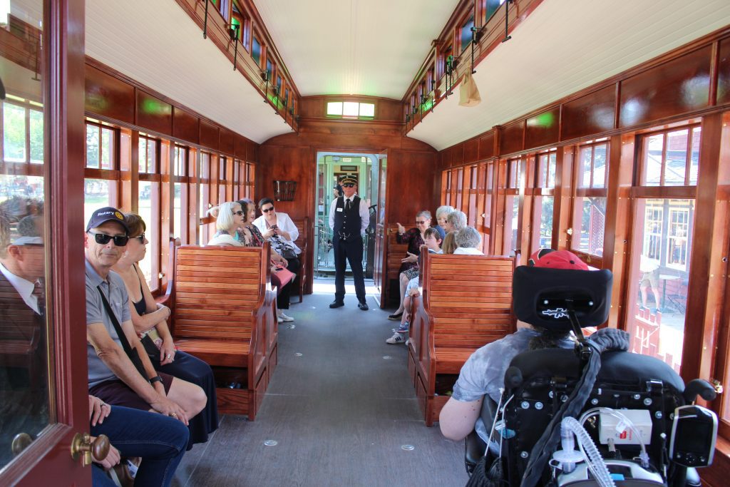 A conductor standing inside a turn of the century styled accessible railcar, with passengers sitting in the seats.