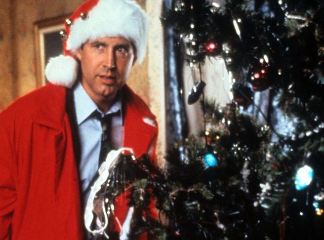 Chevy Chase dressed in a santa suit, peeking behind a christmas tree